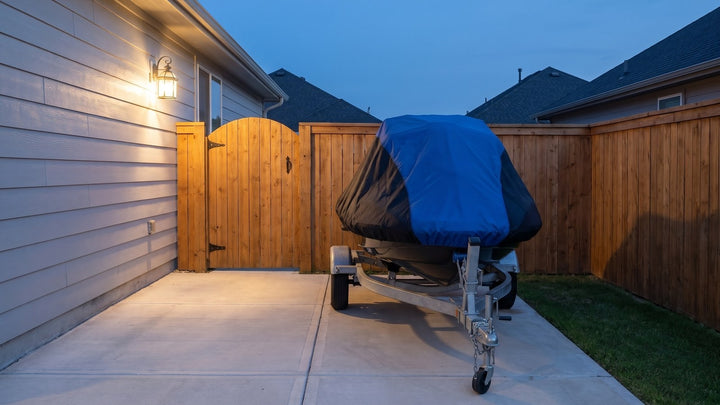 Jet ski on its trailer in a fenced side yard at dusk under a blue and black AllGuard Weatherproof Shield cover
