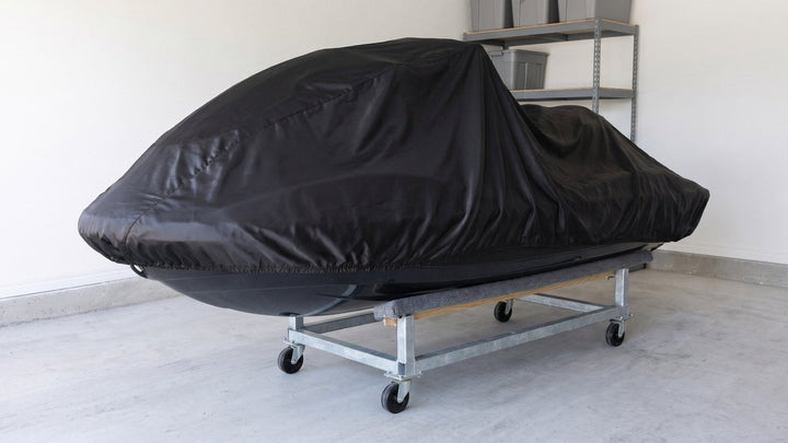 Jet ski under a black AllGuard Satin Shield indoor cover, resting on a wheeled dolly with carpeted bunks in a garage