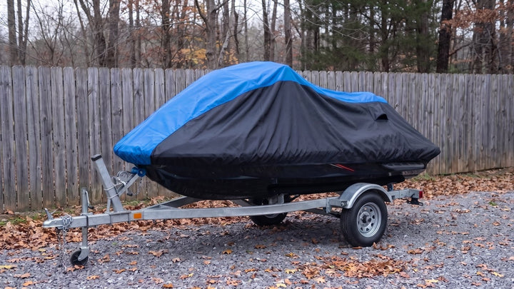 Jet ski on its trailer in a gravel side yard in late autumn, under a blue and black AllGuard Weatherproof Shield cover