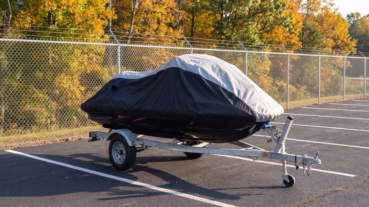 Jet ski on its trailer in a marked space at a fenced outdoor storage lot, under a gray and black AllGuard Weatherproof Shield cover