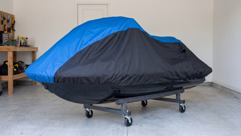 Jet ski under a blue and black AllGuard Weatherproof Shield cover, resting on a four-caster dolly with carpeted bunks in a garage