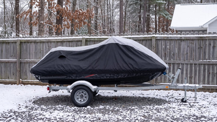 Jet ski on a galvanized trailer in a snowy fenced yard under a gray and black AllGuard Weatherproof Shield cover