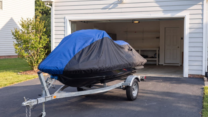 Jet ski on a single galvanized trailer in a driveway in front of an open garage, under a blue and black AllGuard Weatherproof Shield cover