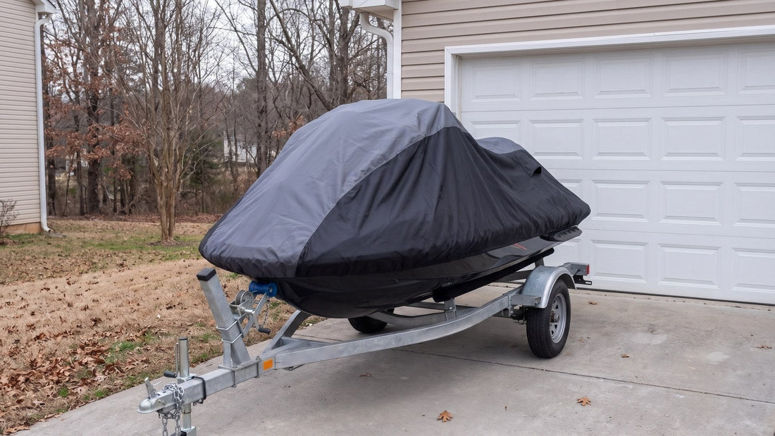 Jet ski on its trailer in a driveway beside a closed garage door on a gray late-winter day, under a gray and black AllGuard Weatherproof Shield cover